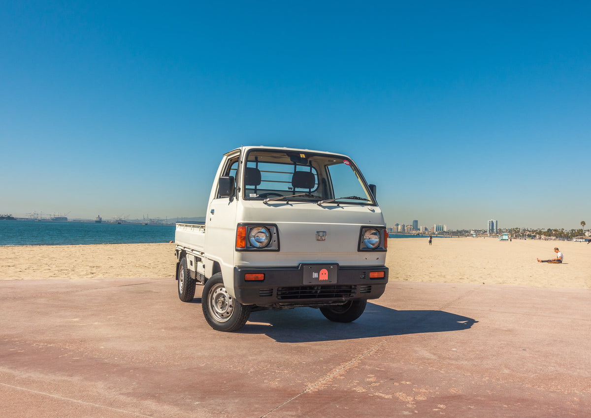 White Honda Acty Kei Truck parked on a sunny beach, showcasing its compact size, classic boxy design, and urban versatility, ideal for enthusiasts and collectors.