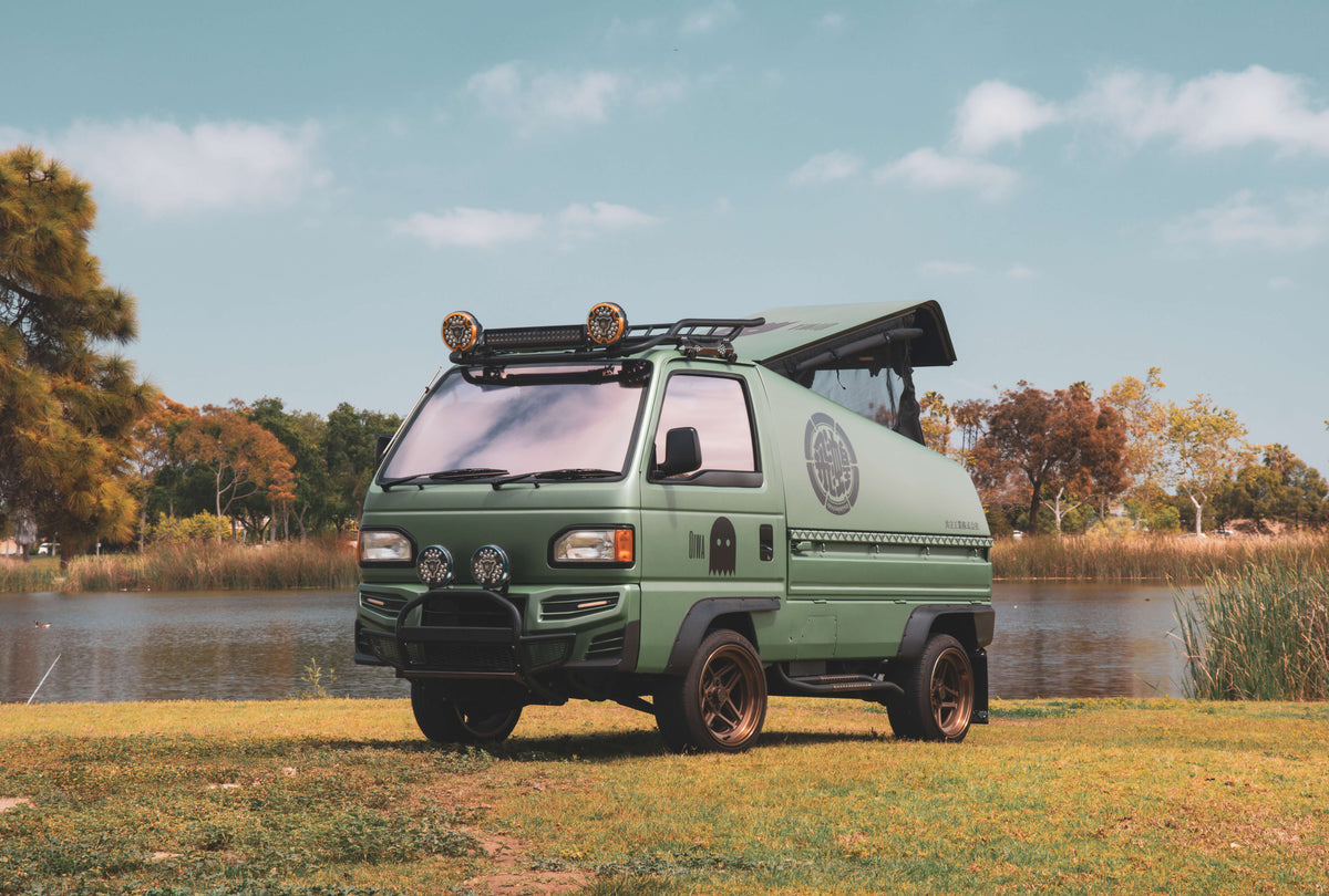 Green Honda Acty Grasshopper kei truck camper with pop-up metal camper shell, lifted suspension, roof rack, and off-road lighting parked in scenic outdoor setting.